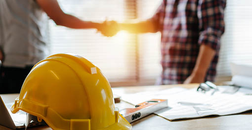 yellow safety helmet on workplace desk with construction worker team hands shaking greeting start up plan new project contract in office center at construction site, partnership and contractor concept